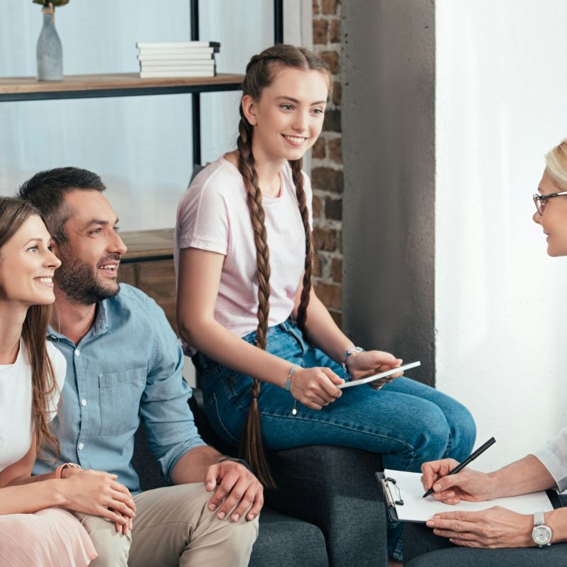 female counselor writing in clipboard on therapy session of family with daughter in office